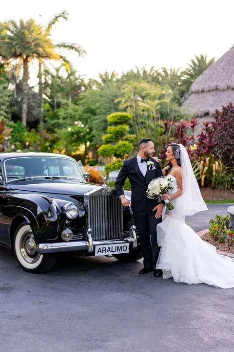 Newlyweds pose beside a black vintage car in the garden.