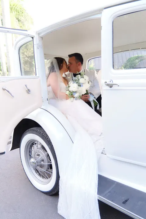 Bride stands beside a white vintage car holding a cascading bouquet.