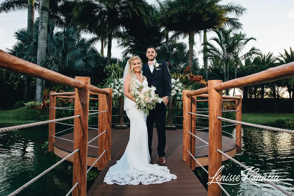 Bride and groom standing on the wooden bridge over the pond with palms and floral arrangements behind them