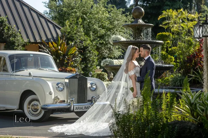 Bride and groom pose beside a silver vintage car and fountain.